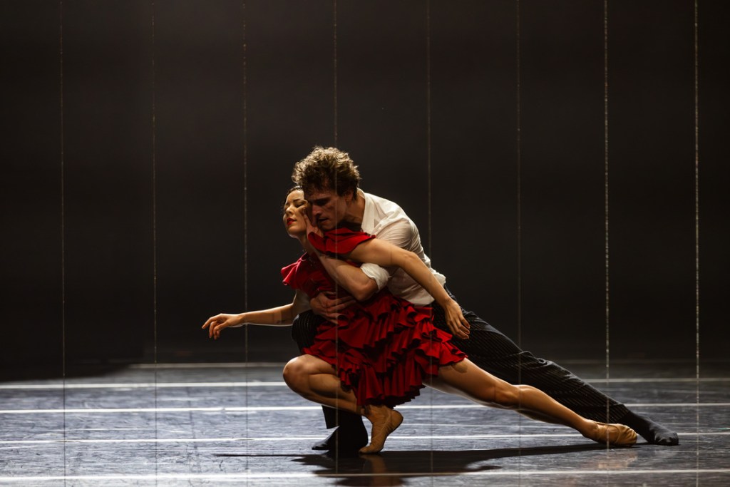 Jill Ogai as Carmen and Callum Linnane as Don José in Johan Inger's 2024 production of Carmen with The Australian Ballet. Ogai, in a striking red dress, confronts Linnane, dressed in a white shirt and dark trousers, in a tense moment that captures the volatile dynamics of their relationship. The minimalist stage design and dramatic lighting underscore the emotional intensity of the scene.