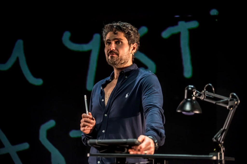 Performer Javaad Alipoor stands on a dimly lit stage, holding a microphone, with digital code and abstract visuals projected behind him. The scene is atmospheric, blending live performance with multimedia elements from Things Hidden Since the Foundation of the World.