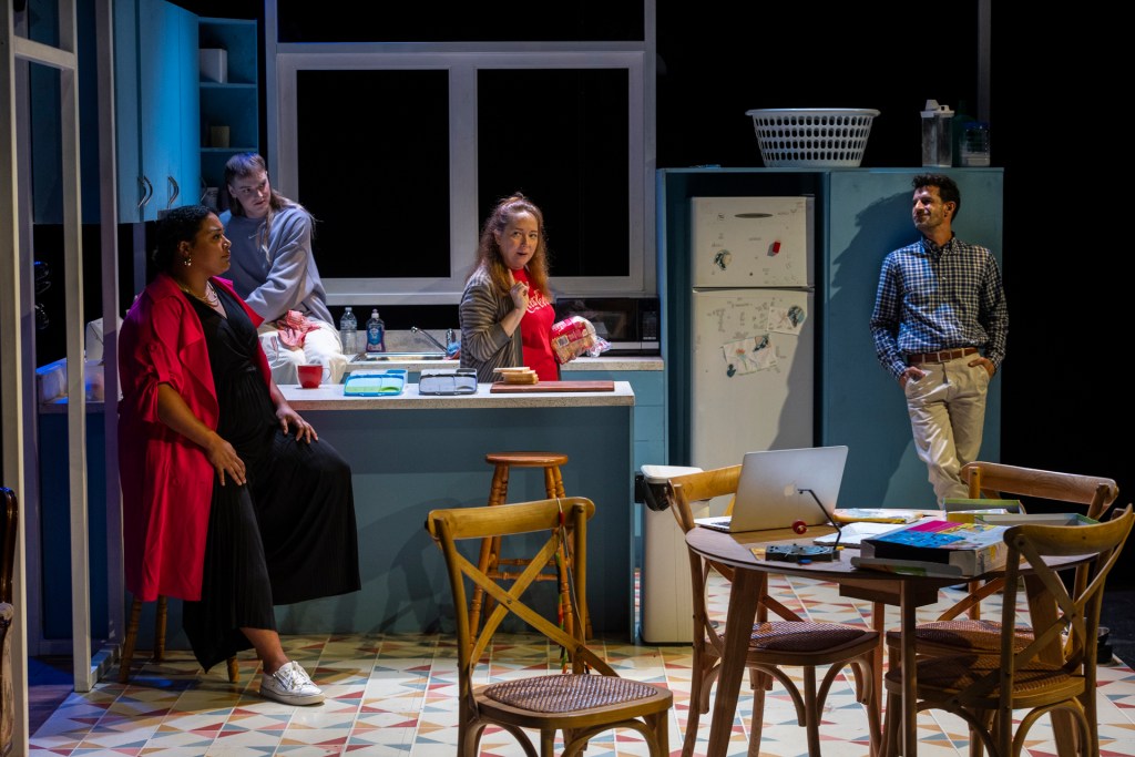 A woman stands center stage, captured mid-expression with arms slightly extended. She wears a Coca-Cola logo tee under a striped cardigan, her long hair flowing past her shoulders. The setting suggests a cozy kitchen living space—softly lit. To her right, man leans near the fridge—in casual attire—observes her, adding a warm, conversational dynamic to the scene. Another man and woman are to the left of her. It seems that the group are conversing. 