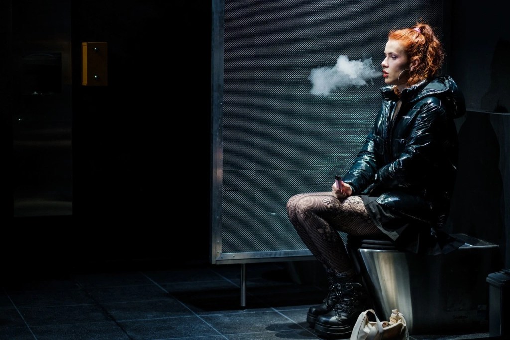 A performer sits alone on a toilet within a stark, all-white bathroom set, lit by a single overhead light. Her posture is introspective, conveying a quiet, reflective moment in Overflow. The minimalist setting draws focus to the emotional weight of the scene.