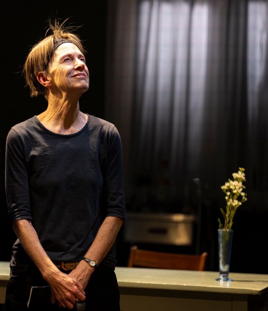 Judy Davis as Helen, standing alone on a minimalist stage with frosted glass windows and bare wooden floors, conveying a moment of introspection.