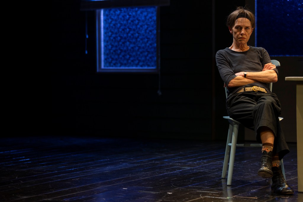 Judy Davis as Helen, sitting alone on a minimalist stage with frosted glass windows and bare wooden floors. Arms crossed in anger and frustration.