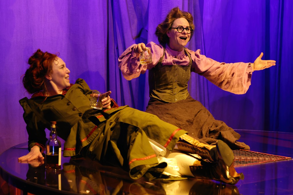 Marie and Hertha sit on the floor, legs outstretched, sharing a bottle of whiskey and laughter, illustrating a moment of levity amidst adversity.