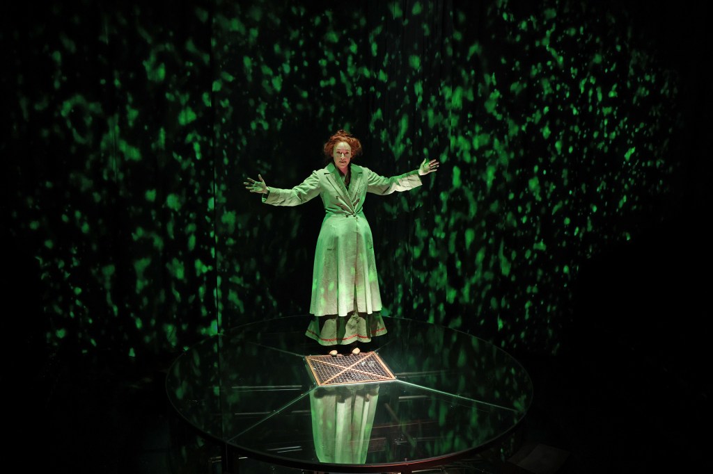A slow-motion sequence depicts Marie Curie appearing to float in the sea, enhanced by ethereal video projections and atmospheric lighting.