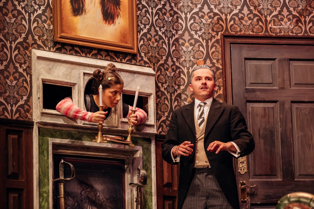 Actors performing a scene from The Play That Goes Wrong, with props in disarray and crew members inadvertently visible on stage