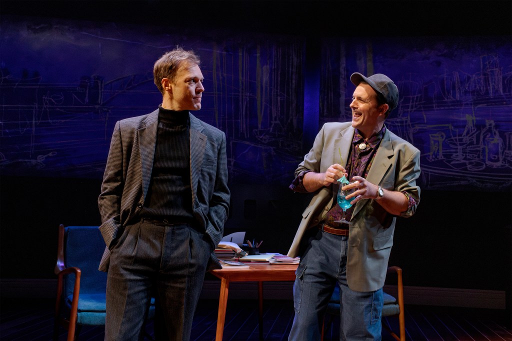 An intense interplay as one actor in formal attire stands pensively beside a desk, while the other, in relaxed clothes and a cap, laughs and stirs a brightly coloured drink, framed by moody lighting and minimalist set design.