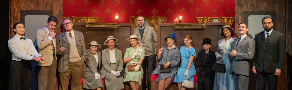 The full cast and crew of The Lady Vanishes at the Genesian Theatre, 2025. The ensemble is captured in a dynamic group shot, showcasing the diverse range of characters and the collaborative effort behind the production. The actors are dressed in period-appropriate costumes, reflecting the 1930s setting of the play.