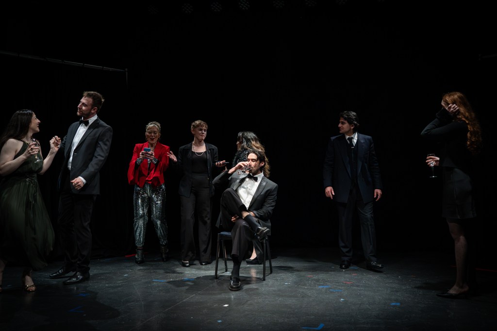 A group of seven individuals dressed in formal attire, engaged in conversation and socializing. One person sits on a chair in the center, while others stand nearby, holding drinks and interacting with each other in a dimly lit stage setting.