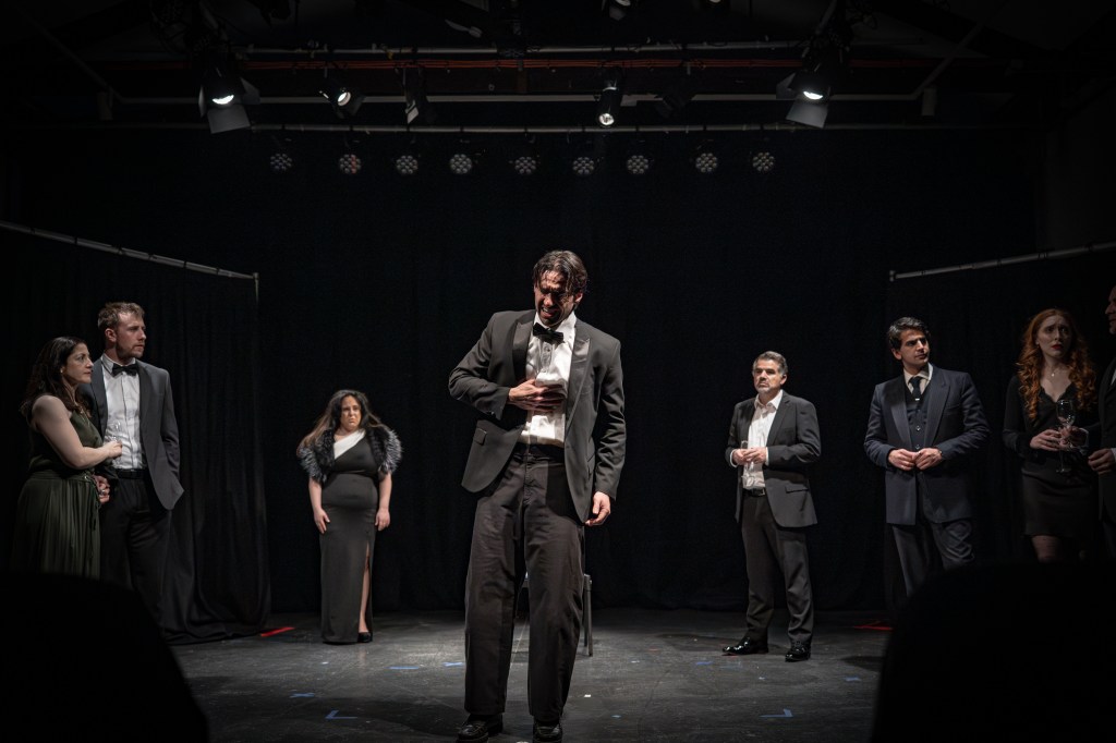 A group of six actors dressed in formal attire performing on stage, with dramatic lighting and a black backdrop.