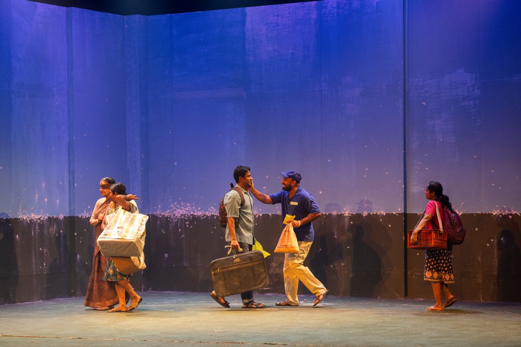 A group of four actors on stage, engaged in conversation while carrying various bags and containers, set against a blue backdrop.