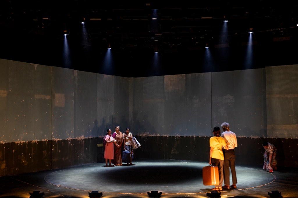 A theater stage with six performers, two men and four women, interacting in a dramatic scene. The backdrop features a large, dark wall with faint lighting effects, and the stage is circular with a spotlight effect.