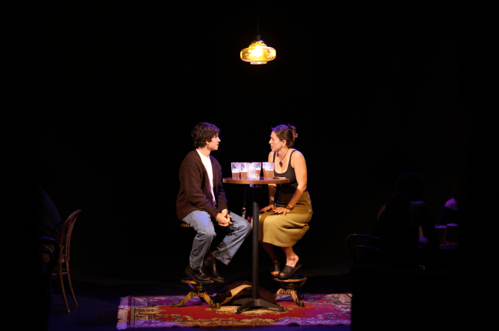 A man and a woman seated at a small round table with drinks, engaged in conversation, in a dimly lit setting. The background features additional blurred figures and a decorative rug under the table.