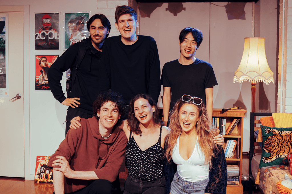 A group of six smiling people posing together in a cozy indoor setting with movie posters on the walls, including a vintage lamp and a bookshelf in the background.