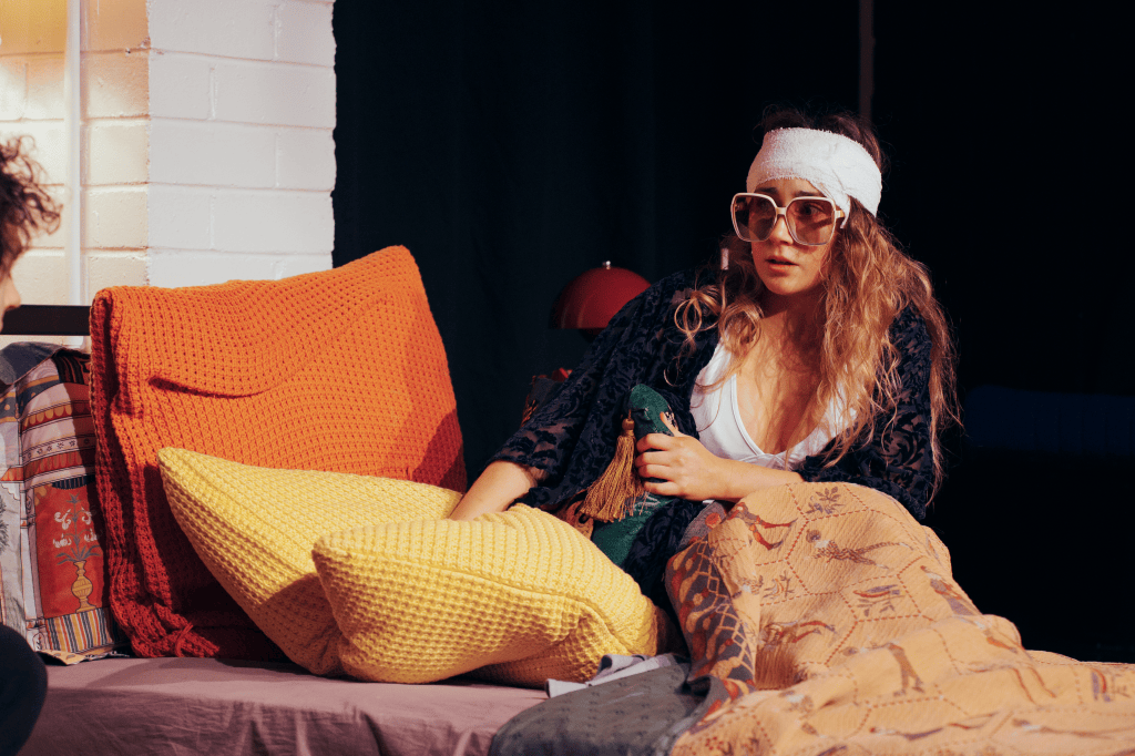 A woman with long curly hair, wearing oversized sunglasses and a headband, sits on a couch surrounded by colorful pillows and blankets, appearing relaxed or contemplative.