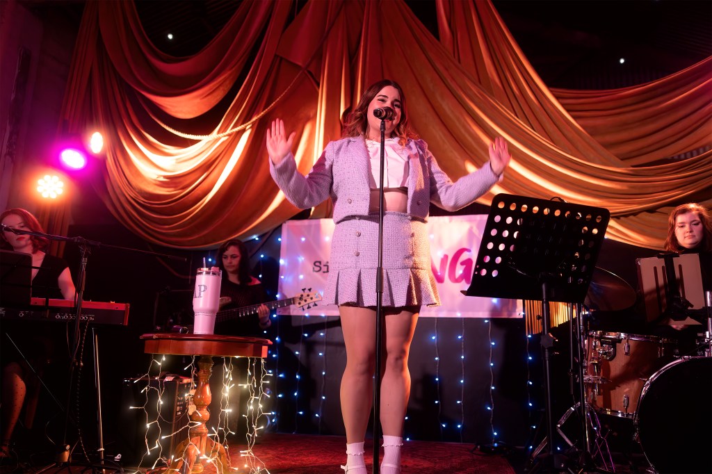 A female singer performs on stage with a microphone, wearing a stylish outfit, surrounded by musicians on keyboards and drums, with colorful stage lighting and decorative drapery in the background.