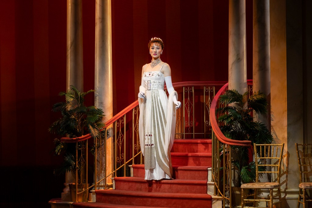 A performer in an elegant, white gown stands on a staircase, showcasing a classic and sophisticated look in a theatrical setting.