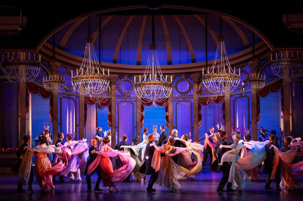 A vibrant musical dance scene on stage featuring numerous performers in colorful costumes, surrounded by elegant chandeliers and a grand set design.