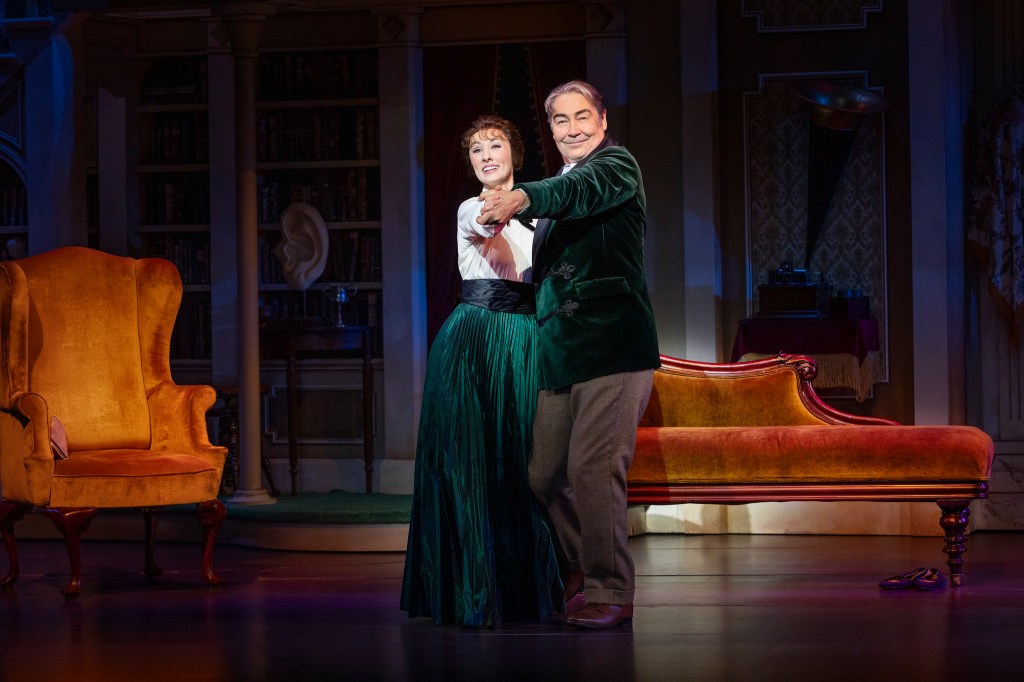 A man and a woman dance together on stage, surrounded by a vintage interior with a large armchair and a sofa. The woman wears a teal skirt and a white top, while the man is dressed in a green velvet jacket, both smiling as they enjoy the moment.