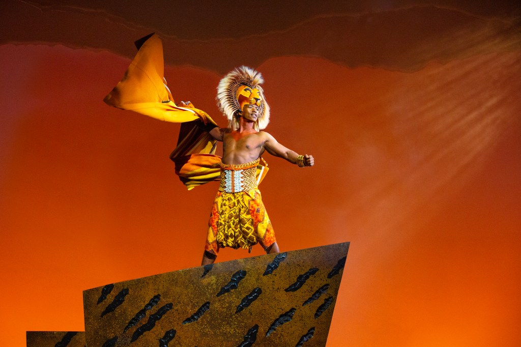 A performer dressed in a vibrant yellow and orange costume, posing dramatically on a set piece against a warm orange background, evoking themes of power and identity.