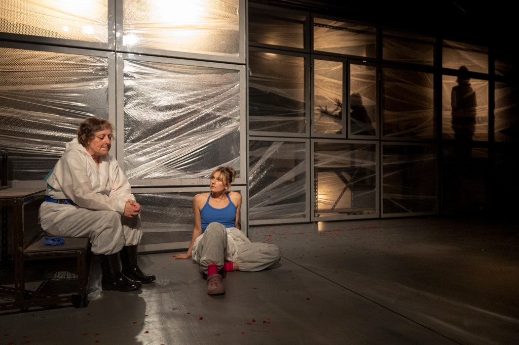 Two women seated in a dimly lit space, one in a white protective suit and the other in a blue tank top and light pants, with plastic wrap covering the backdrop. Shadows of figures are visible in the background.