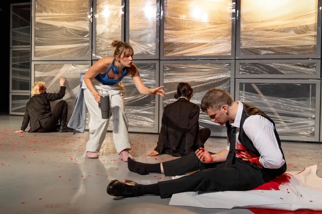 A dramatic scene from a theatrical performance showing actors in various states of distress. A woman in a blue top gestures towards a man in a suit, who is sitting on the floor with blood on his hands. Other actors are positioned in the background, some engaged with the set and others seemingly in turmoil.