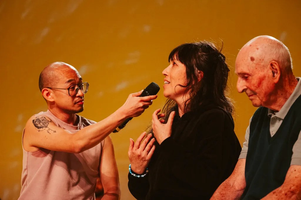 A group of three people engaged in conversation on stage, with one person holding a microphone. The setting features a vibrant yellow background.