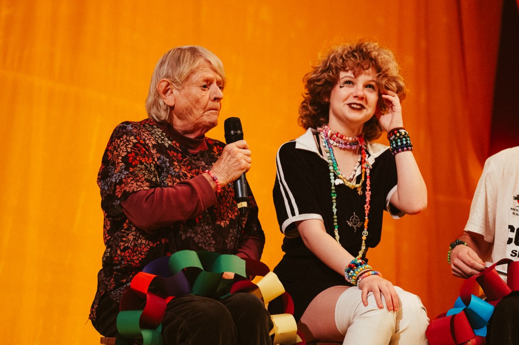 An elderly person with gray hair sitting on a stage holding a microphone, wearing a floral shirt and bracelets, alongside a young person with curly, blonde hair dressed in a black and white outfit, adorned with colorful necklaces and bracelets, both smiling against a vibrant orange backdrop.